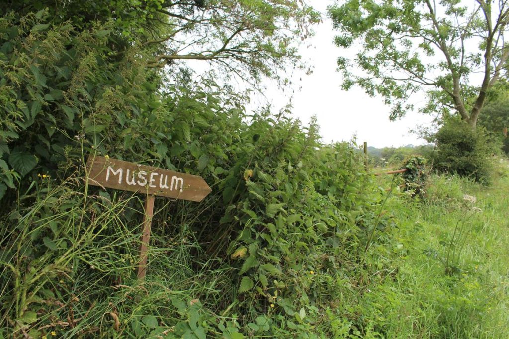 A wooden hand-painted sign for a museum standing in a bank of nettles