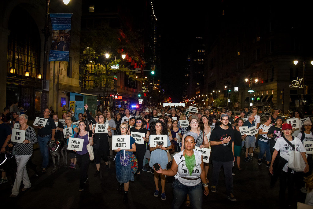 A large parade of people on a night-time street in Philadelphia. They carry placards bearing people's names.