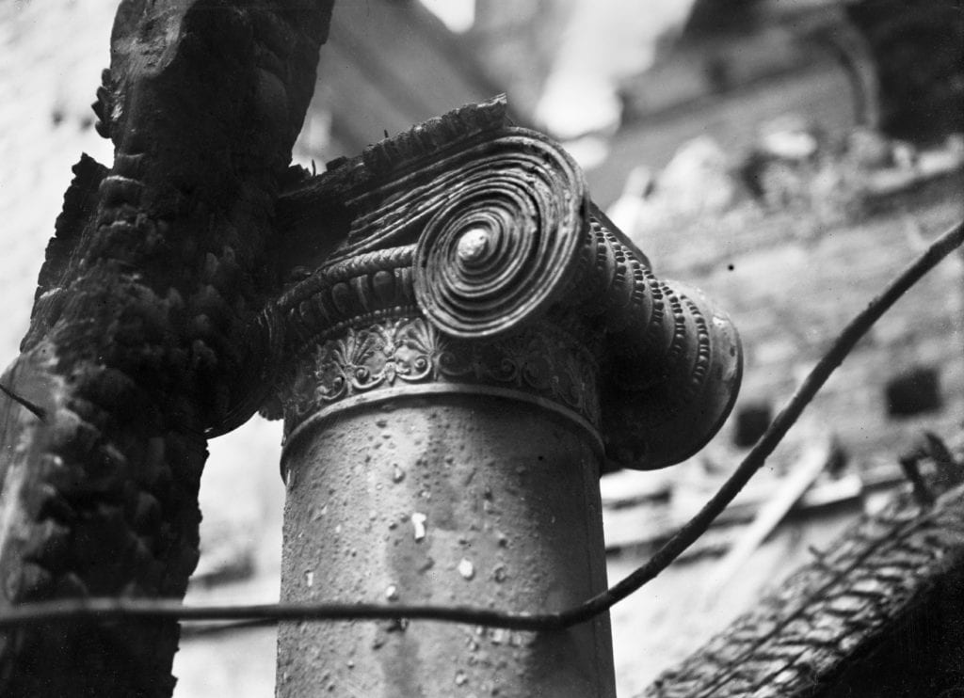 A damaged Ionic column capital. Charred wood leans against it and more is visible in the background. A loose wire runs across the bottom of the image.