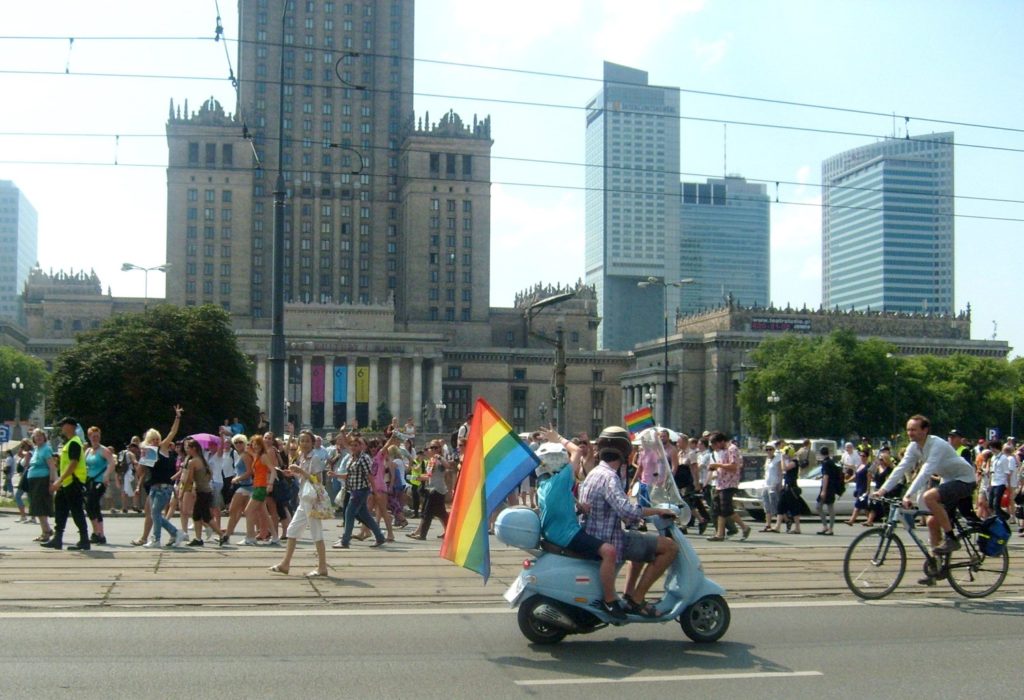 A couple on a scooter holding rainbow flags drive past a busy street scene